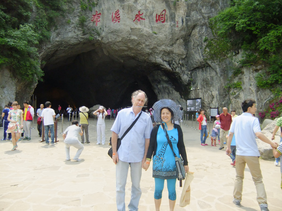 A couple posing in front of a large cave entrance with Chinese characters carved in red on the rock face above, surrounded by other tourists at a scenic outdoor site.