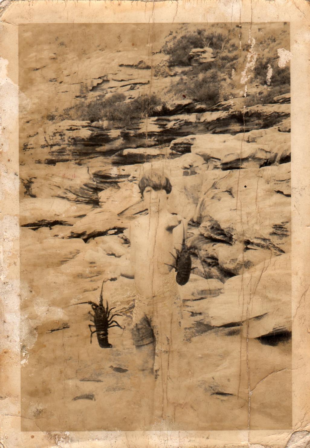 Vintage sepia-toned photograph or print depicting rocky desert cliffs with small dark insect-like silhouettes near the lower left and center, surrounded by aged tan paper borders and visible wear, scratches, and discoloration.