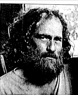 A black and white close-up portrait of a man with long curly hair, a full beard, and a serious expression, shown in a grainy, high-contrast print style.