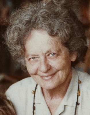A close-up portrait of an elderly woman with curly gray hair, smiling and wearing a light-colored collared shirt and a beaded necklace.

