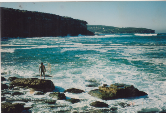 A scenic coastal photograph showing turquoise ocean waves crashing around dark rocks, with a person standing on a rock in the foreground and a large cliff or headland in the background under a pale sky.

