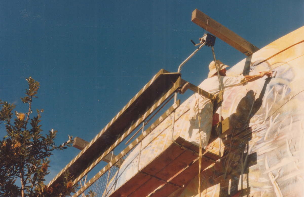 A person in white clothing works on a large mural or wall under a wooden support structure, with scaffolding and a clear blue sky in the background.


